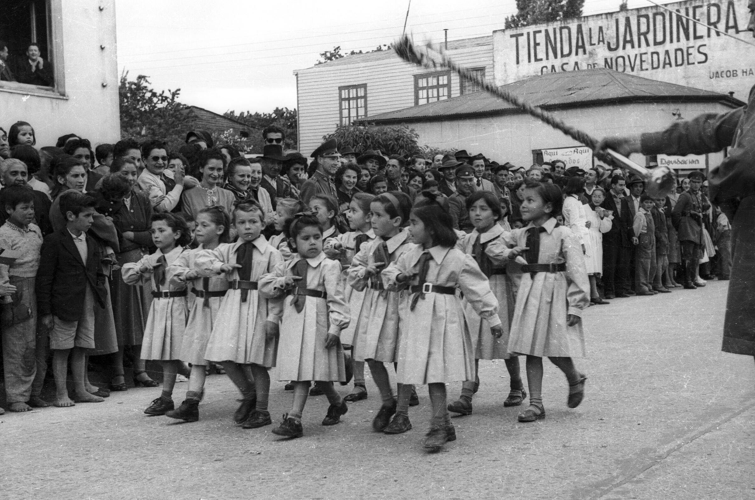 Plano entero de un grupo de niñas, pertenecientes a una brigada no identificada, desfilando formadas en dos hileras mientras realizan un saludo al pecho. En un costado un grupo numeroso de personas las observa avanzar desde el frontis de la municipalidad