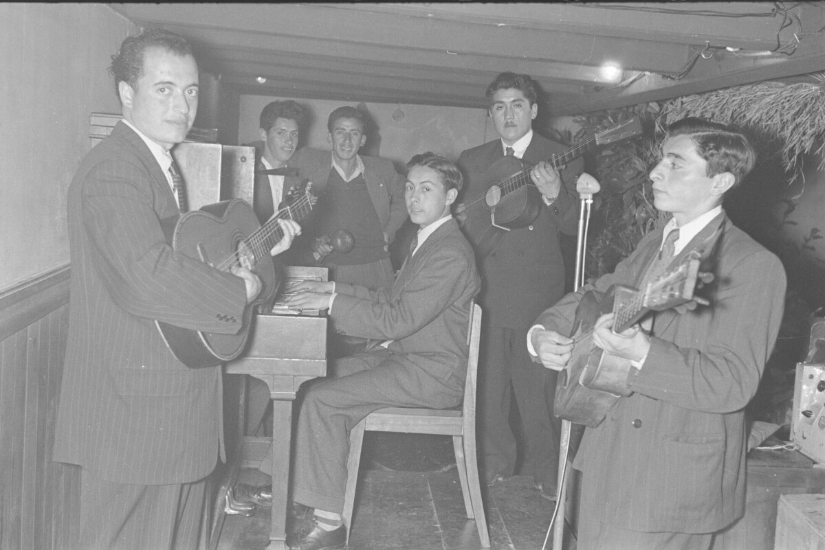 plano general de un grupo musical tocando sus instrumentos. Algunos de ellos están mirando hacia la cámara. Todos visten formalmente. Se encuentran tocando en el interior de un local no identificado, durante la celebración de una coronación a reina no identificada