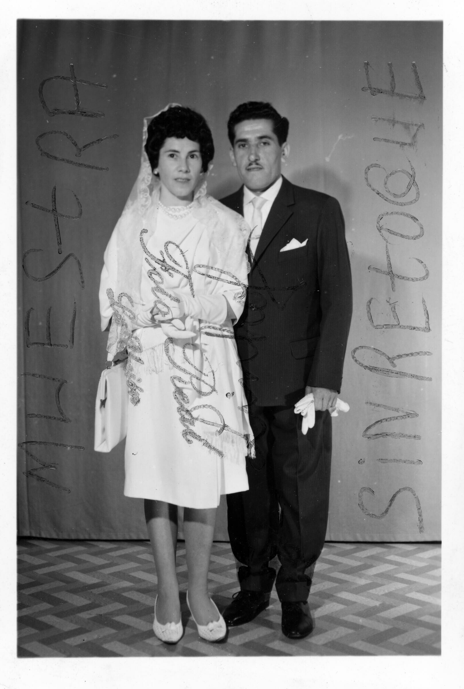 Retrato de estudio de Plano entero de Omar Peralta y so novia Herminda, posando tomados del brazo. La novia lleva colgando una cartera y el novio tiene en la mano sus guantes