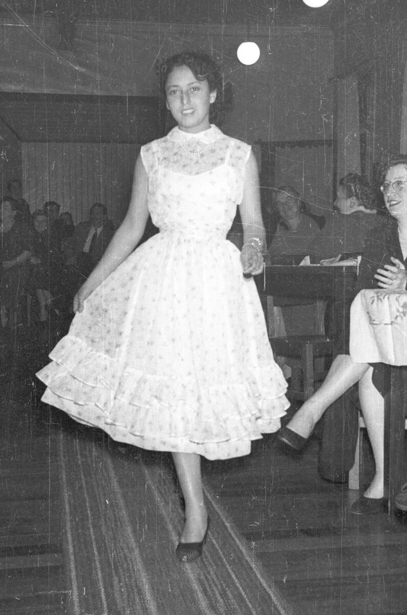 Plano entero de una mujer modelando un vestido mientras camina por una alfombra. En un costado hay una mujer sentada junto a una mesa que la está observando junto a otras personas las cuales se encuentran en el fondo del local donde se está llevando a cabo una exhibición de Vestidos