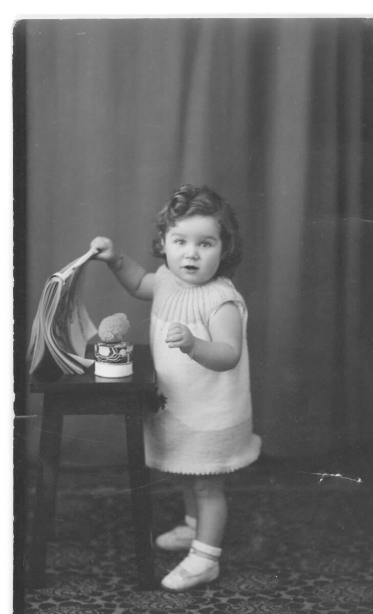 Retrato de estudio de Plano entero de una niña posando parada junto a una mesa, de la cual está sacando una revista. Ella lleva puesto un vestido y se encuentra parada sobre una alfombra