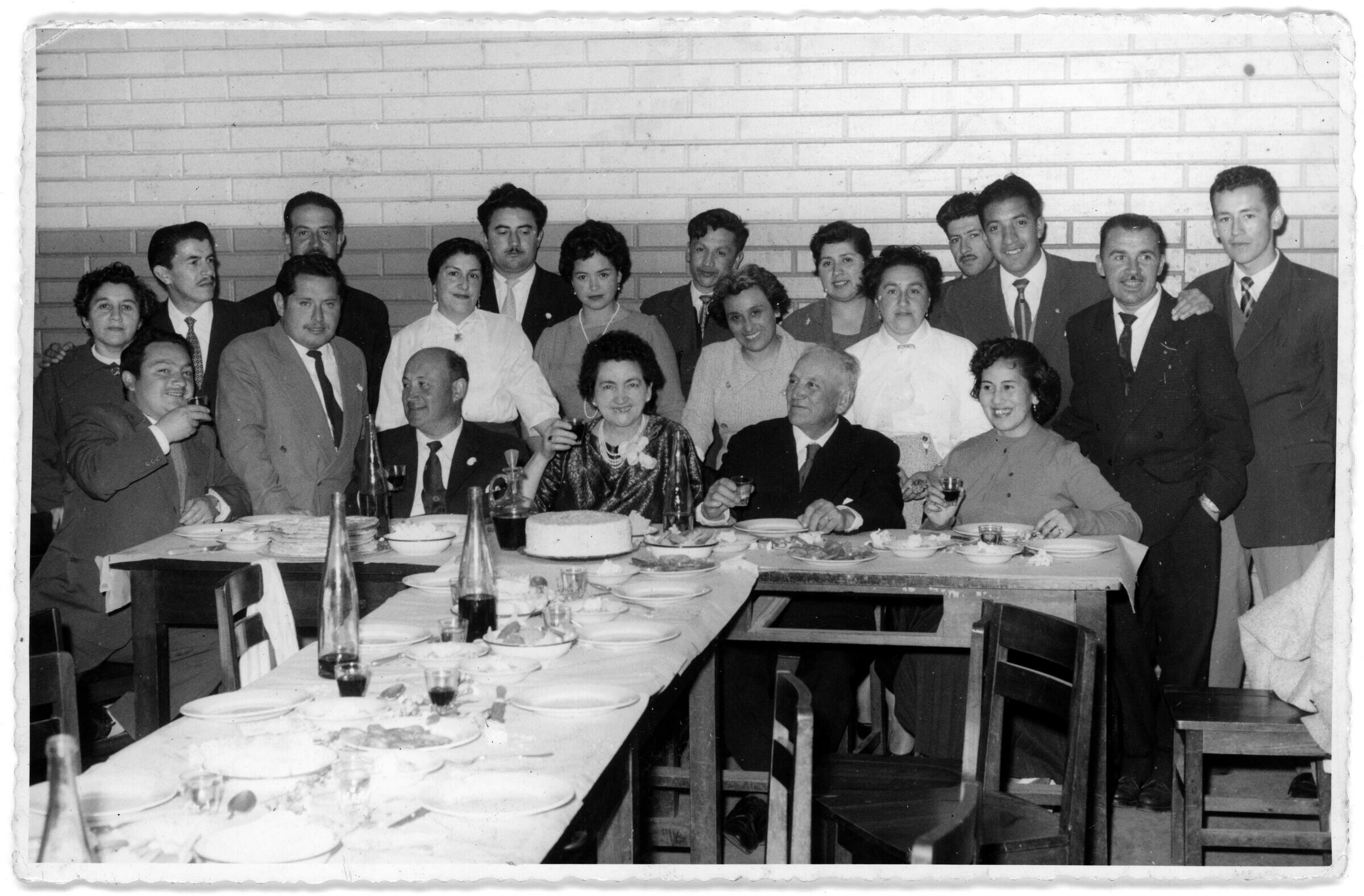 Retrato grupal de plano entero de Hombres y Mujeres posando detrás de una mesa. Solo cuatro personas se encuentran sentados mientras que el resto está posando de pie por detrás de sus sillas. Todos visten formalmente y en la primera hilera están brindando con una copa en la mano. Se encuentran en el interior de una infraestructura no identificada durante una celebración