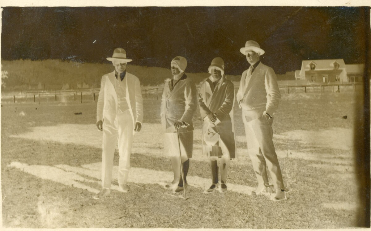 Retrato de plano entero en negativo de dos hombres y dos mujeres posando de pie sobre el pasto. A sus espaldas hay una casa de dos pisos y un cerro poblado de árboles