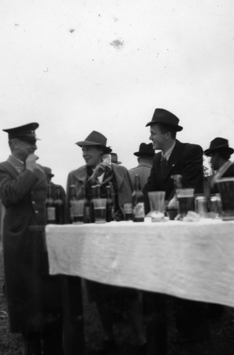 Plano entero de Olga Boettcher, conversando con un carabinero con una copa en la mano. Los acompaña un hombre de Traje y sombrero. Están ubicados al final de una mesa, sobre la cual hay una gran cantidad de botellas y vasos. Se encuentran celebrando en el interior de un recinto no identificado