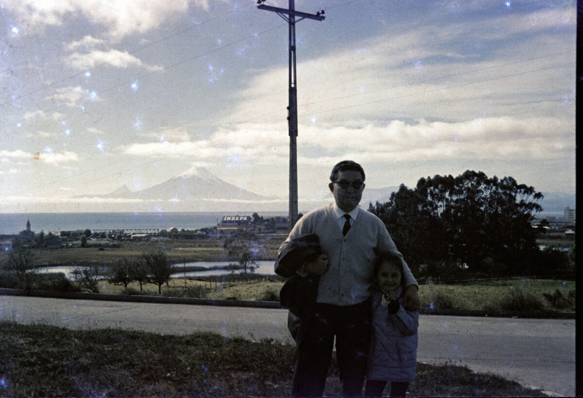 Retrato plano americano de Hombre con Traje abrazando a niña. Detrás de ellos un paisaje con árboles, el lago Llanquihue y los volcanes Osorno y Puntiagudo.