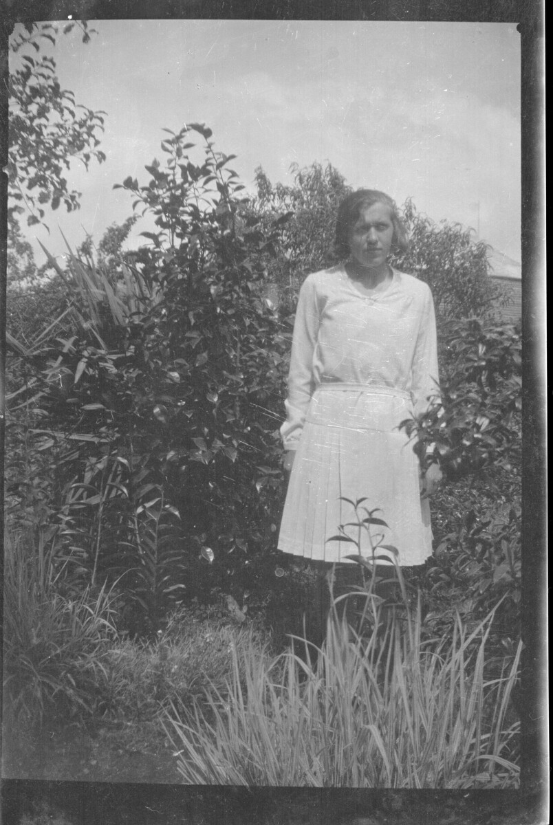 Plano entero de una mujer joven posando de pie entre plantas de un jardín, ella viste una blusa y una falda plisada.