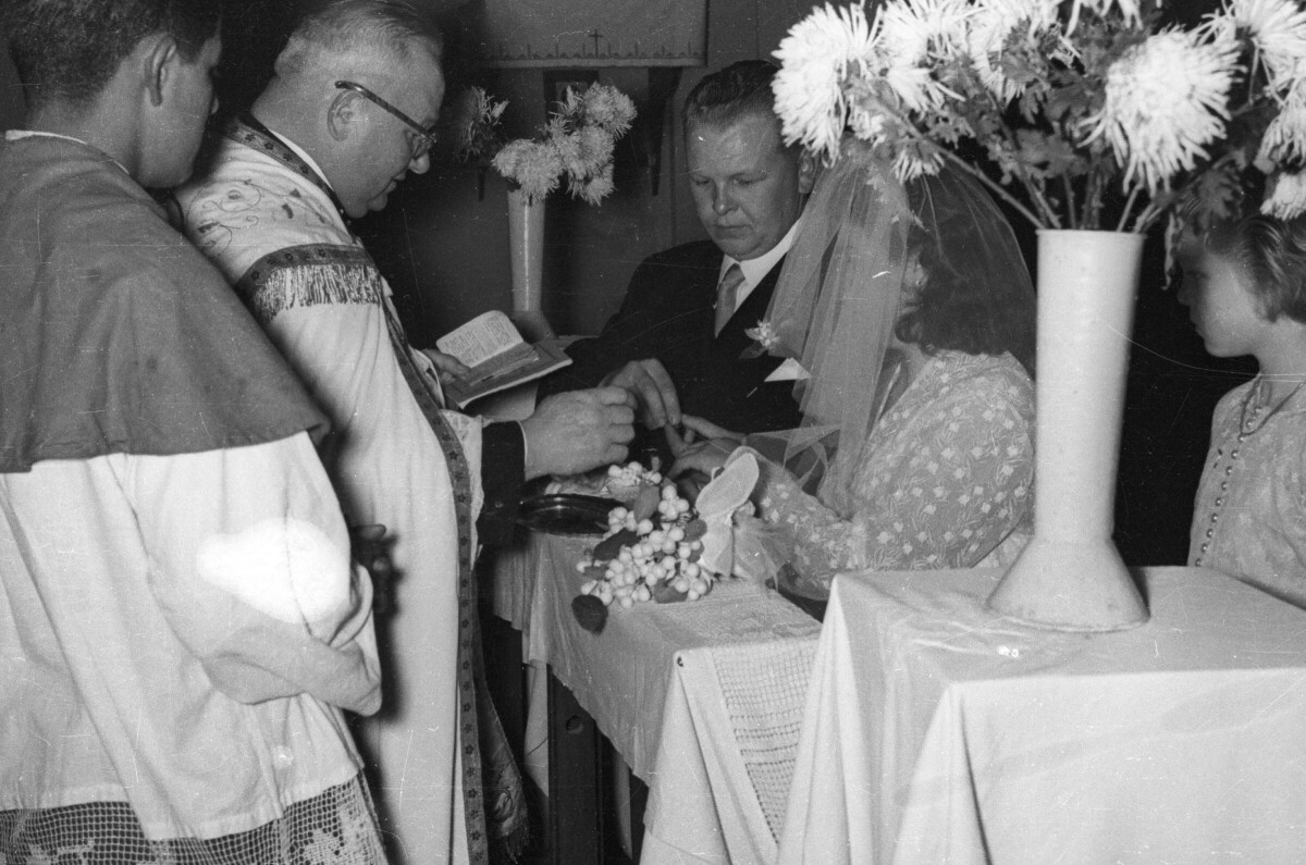 Plano medio largo de un sacerdote entregándole las argollas a una pareja de novios que se encuentran arrodillados en el altar. Los observa uno de sus acólitos y una Niña. Se encuentran en el altar durante la misa de celebración de su matrimonio, realizada en el interior de una parroquia no identificada