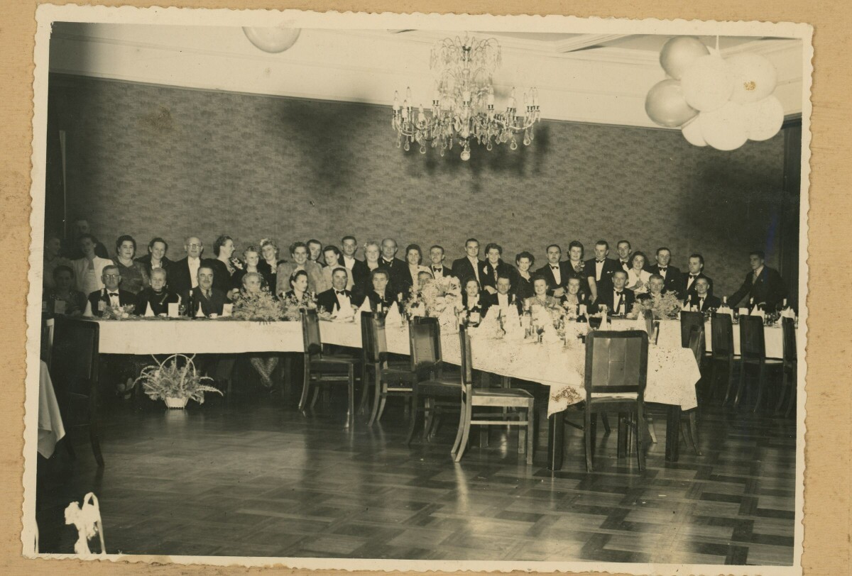 Retrato grupal de plano entero de Mujeres y Hombres formados en dos hileras por detrás de una larga mesa durante un banquete realizado en el interior de una estructura no identificada.