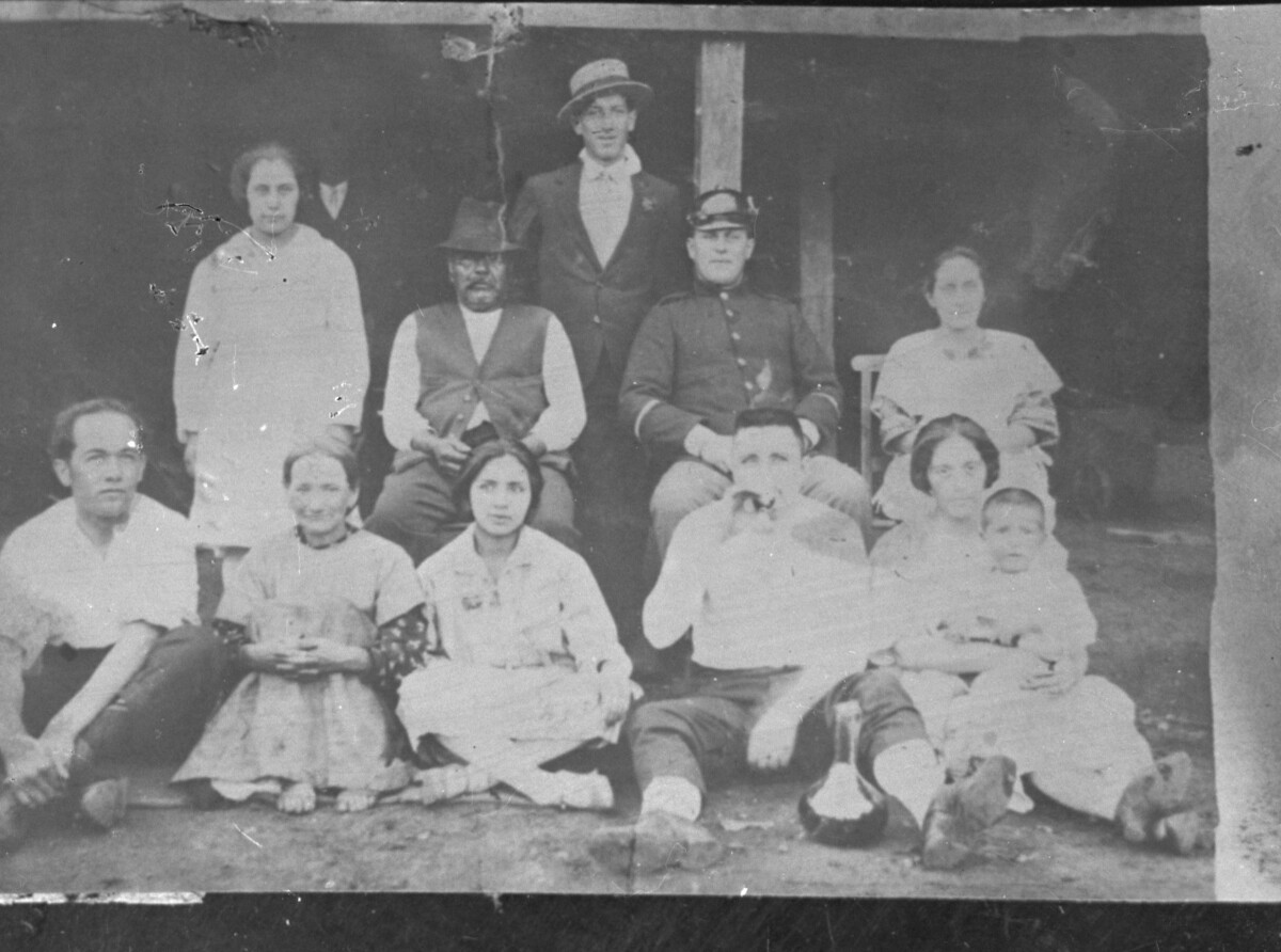 Reproducción de fotografía. Retrato de plano entero de un grupo de Hombres y Mujeres, algunos sentados en sillas y otros sentados en el suelo, todos posando de frente en un lugar no identificado