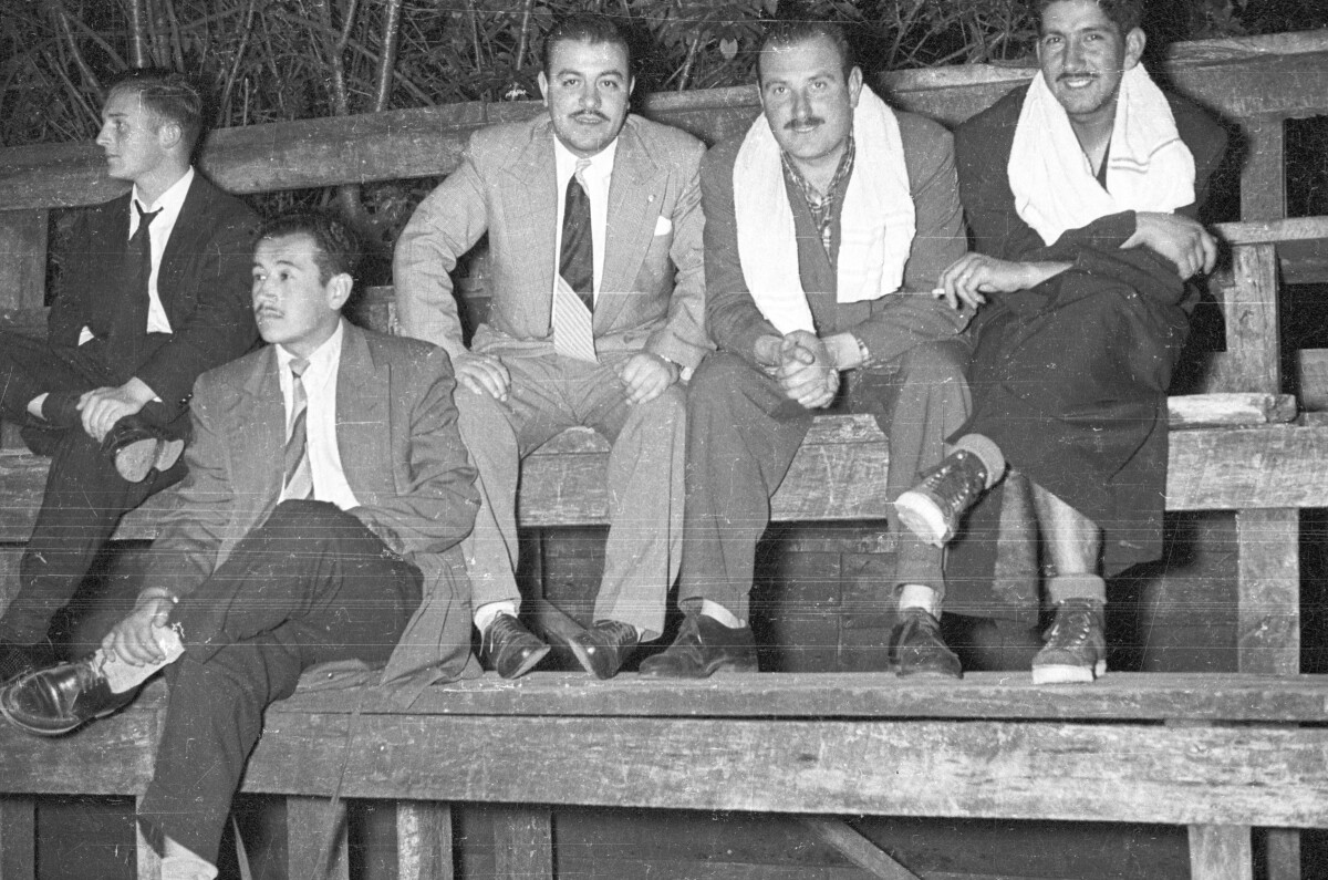 plano general de tres hombres vestidos posando. Dos de ellos viste de traje mientras que el tercero tiene una bata y una toalla en el cuello. Junto a ellos hay otros dos hombres observando el juego sentados en unas graderías durante la noche.