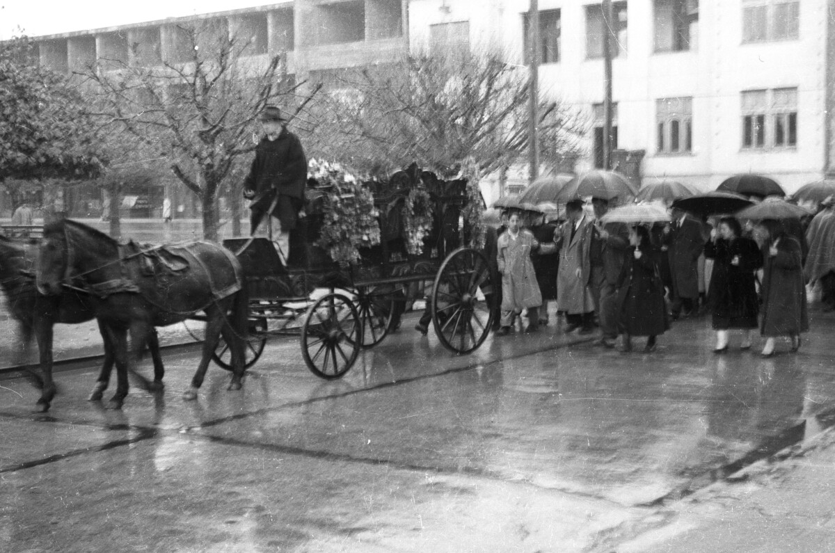 Plano entero de grupo de Hombres con Trajes y abrigos y Mujeres con Vestidos y parkas, algunos con paraguas caminando detrás de una carroza fúnebre tirada por caballos