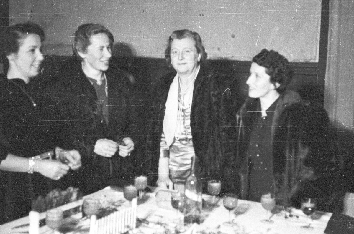 Plano entero de cuatro mujeres conversando de pie detrás de una mesa durante una celebración en el interior de una local