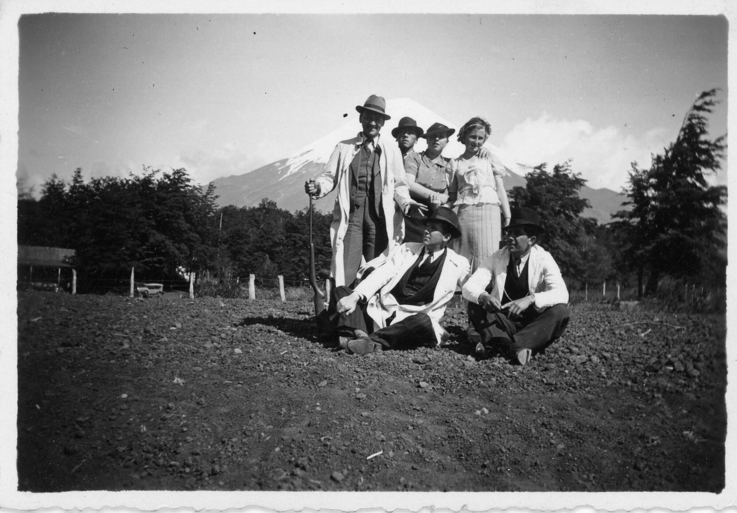Retrato grupal de plano entero de mujeres y hombres posando. El primer hombre parado de izquierda a derecha tiene consigo una escopeta. En el fondo hay un volcán no identificado.