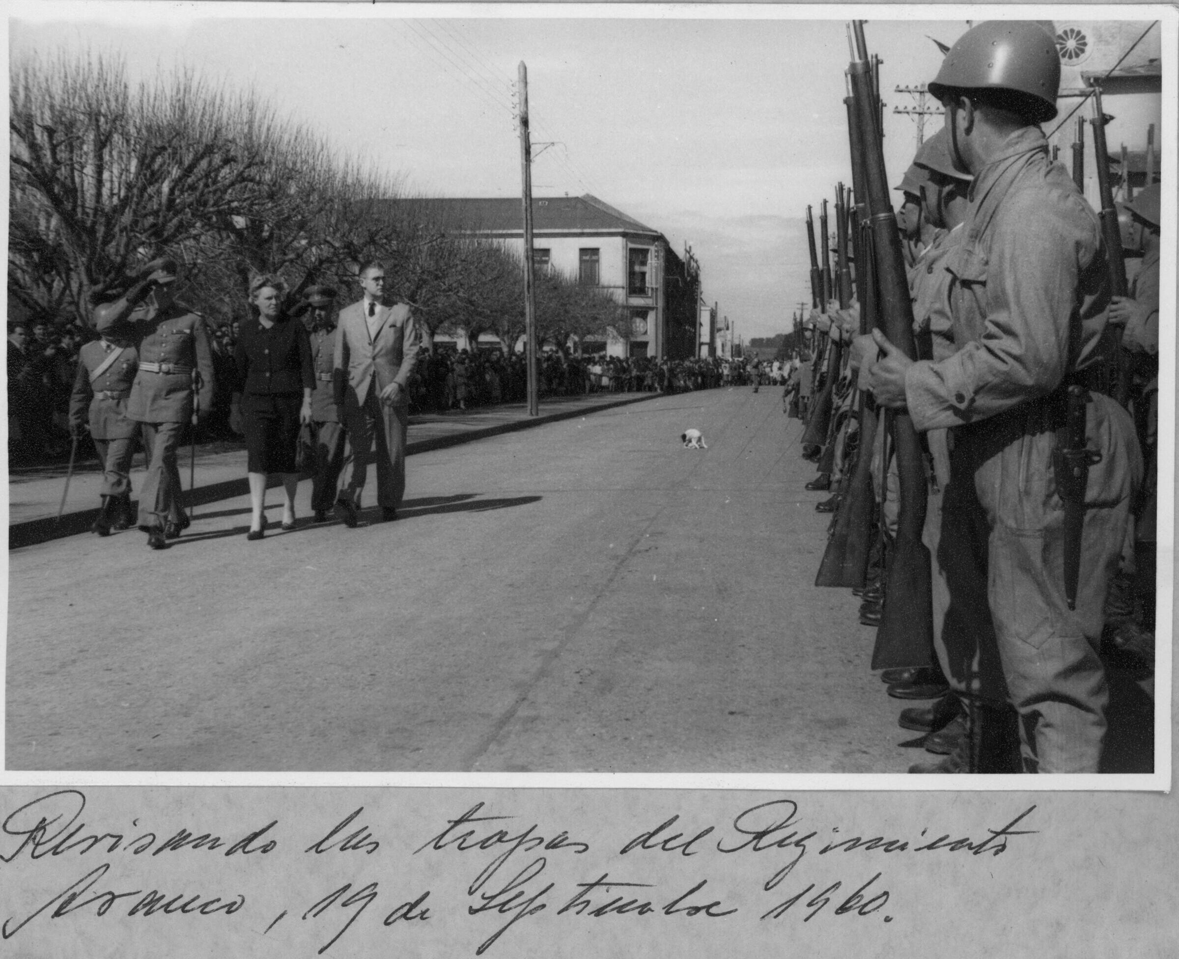 plano general de Olga Boettcher, caminando junto a tres carabineros y un hombre vestido de Traje, mientras estos realizan un saludo a las tropas militares que se encuentran formadas a un costado de la calle con su fusil en la mano. Al medio de la calle, hay un perro parado. A un costado de la plaza de La Concordia, hay un grupo numeroso de personas que se encuentran reunidas observando la actividad