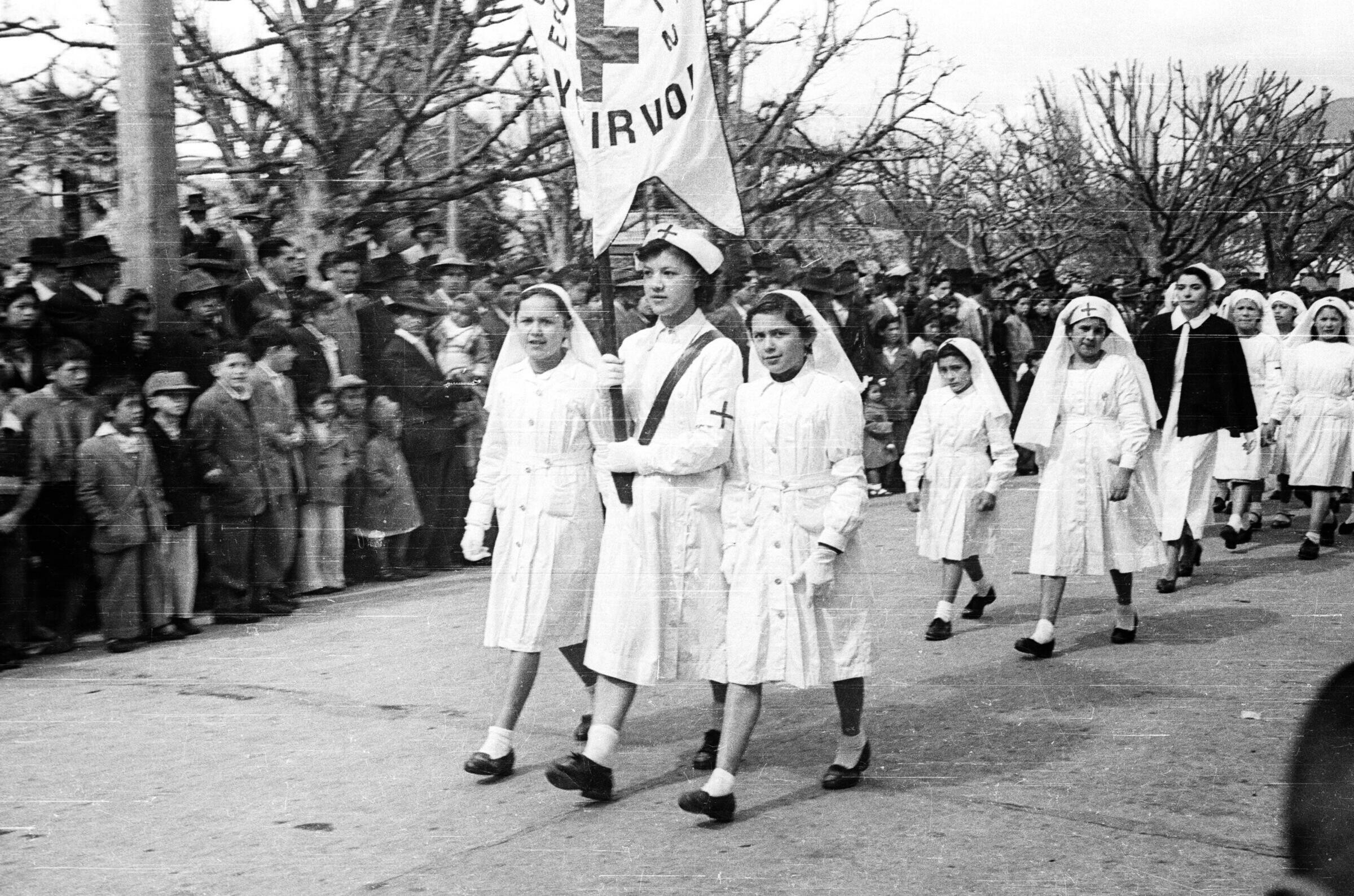 plano general de un grupo de Niñas caminando vestidas de blanco con una túnica en su cabeza que tiene dibujada una cruz sobre la frente. En un comienzo de la fila van tres Niñas, la del medio lleva el pendón de la Cruz Roja Juvenil de la Escuela Nº2. En un costado de la calle hay un grupo de Niños, Niñas, Hombres y Mujeres observándolas de pie sobre la vereda durante un desfile por las calles de La Unión