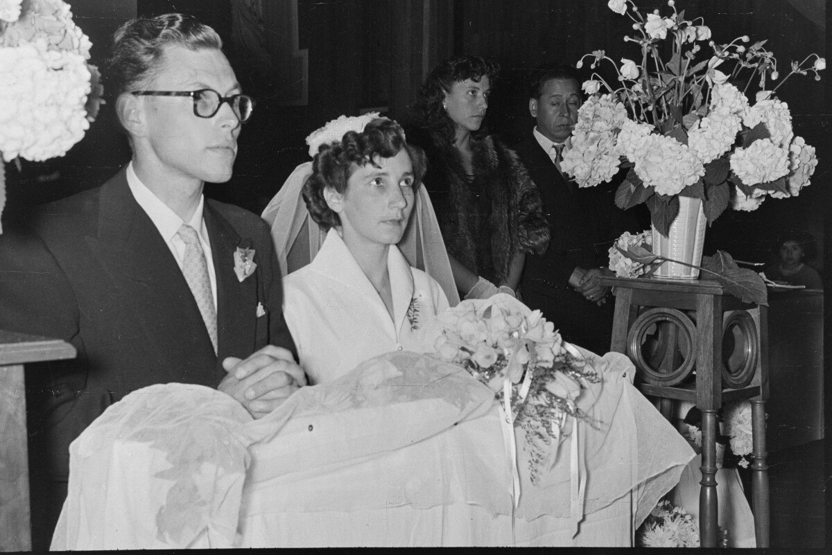 Plano medio largo de una pareja de novios, hincados sobre un reclinatorio el cual está ubicado en el altar. En el fondo, sus padrinos de bodas se encuentran parados durante acompañándolos durante la misa de celebración de su casamiento, realizada en el interior de la parroquia San José