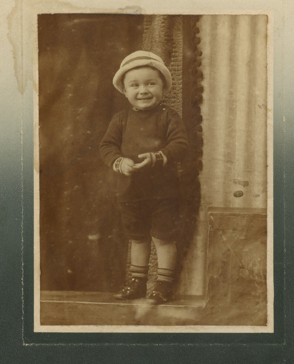Retrato de plano entero de un niño con sombrero, sonriendo mientras posa parado en un peldaño. Lugar no identificado