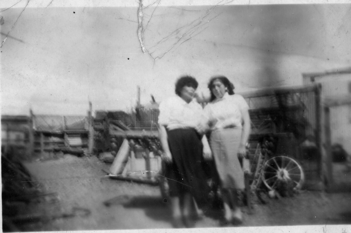 Retrato de plano entero de dos mujeres posando de frente. A sus espaldas, hay algunos objetos no identificados, arrumados a un costado de un cerco de madera.