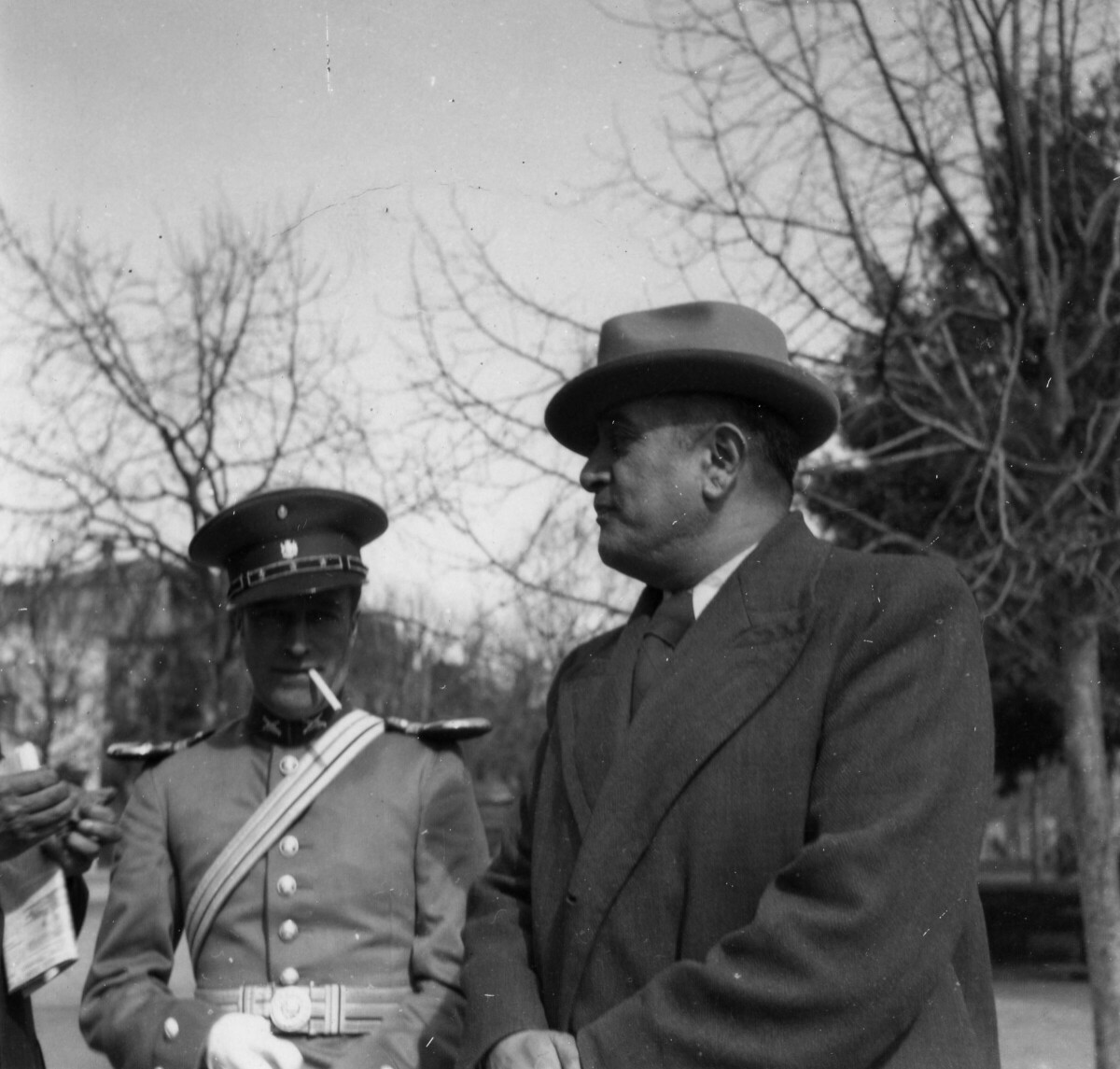 Plano medio largo de un carabinero parado con un cigarro en la boca mientras a su lado, se encuentra un hombre con sombrero acompañándolo. Están parados sobre la calle, a un costado de la plaza de La Concordia