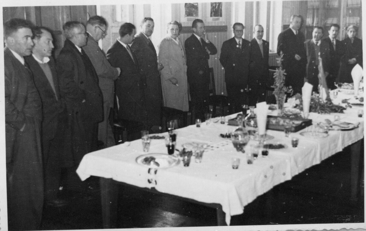 plano general de un grupo de hombres parados detrás de una mesa. En esa misma hilera, se encuentra parada Olga Boettcher, gobernadora de La Unión. Es la primera mujer de izquierda a derecha. Se encuentran en el interior de una infraestructura no identificada durante un banquete