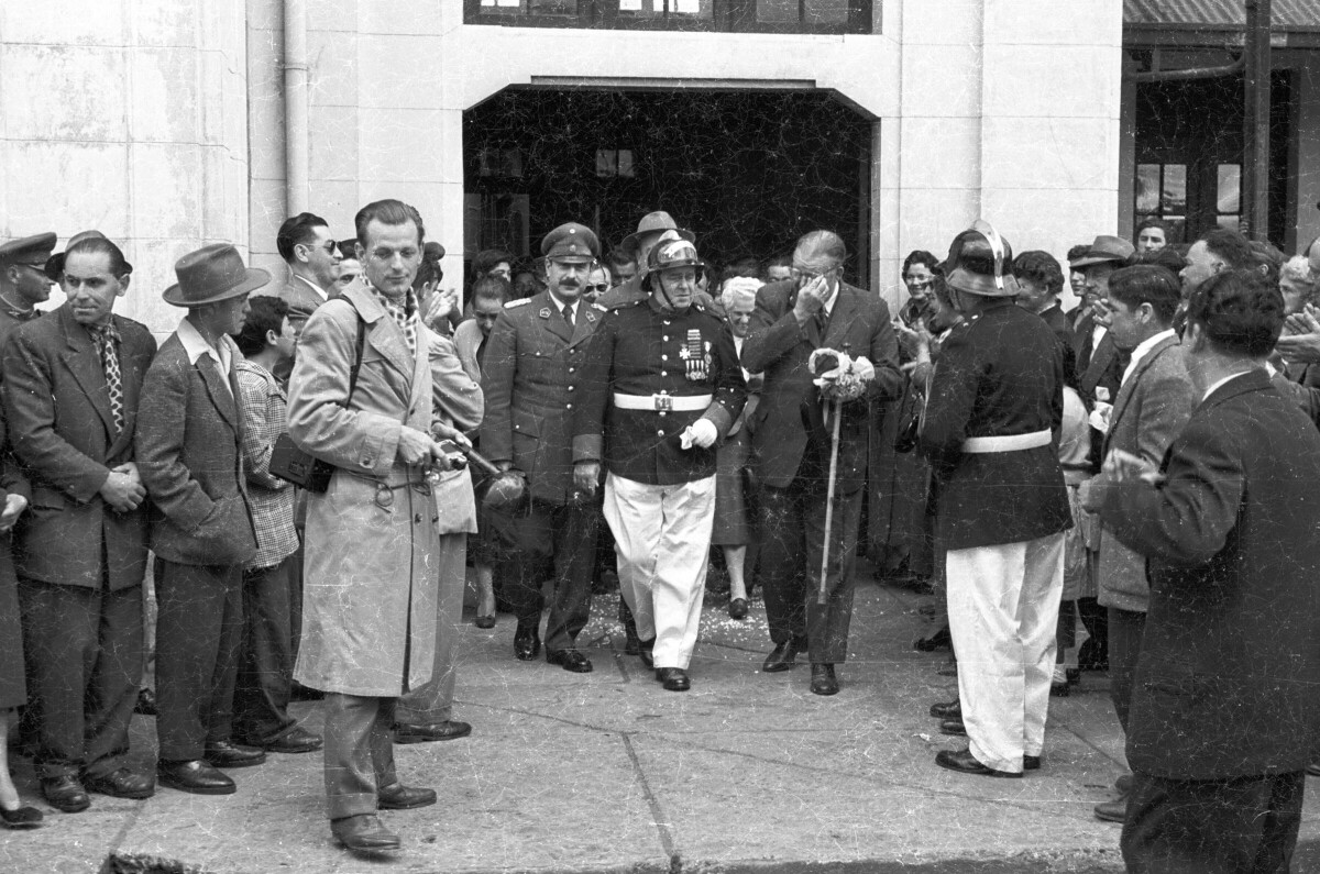 plano general de Hombres vestidos de Traje, reunidos alrededor de un grupo de uniformados y Carlos Vogel, quienes vienen saliendo de la estación de trenes durante la llegada del alcalde a la ciudad de La Unión