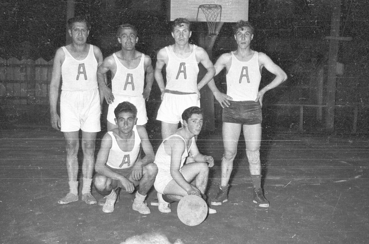 Retrato grupal de plano entero de un equipo masculino de baloncesto posando todos juntos. Algunos están de pie y dos de ellos en cunclillas. En sus poleras llevan escrita la letra A'. están posando sobre la cancha durante la noche.