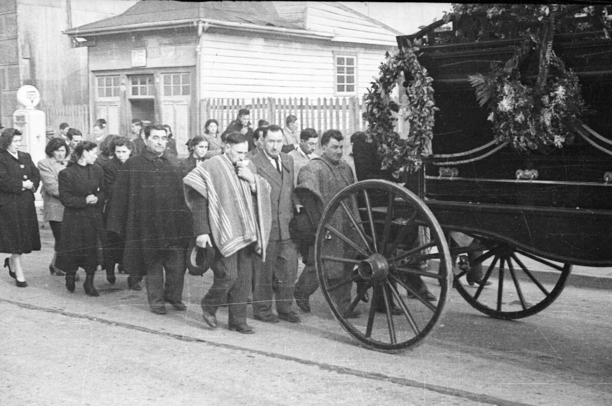 plano general de un grupo de hombres y mujeres siguiendo la carroza fúnebre por una calle a la salida de la iglesia