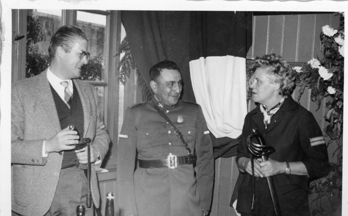 Plano medio largo de Olga Boettcher conversando con un carabinero y un hombre vestido de Traje, mientras sostiene una espada enfundada en manos. Se encuentra en el interior de una infraestructura no identificada durante su visita al lugar