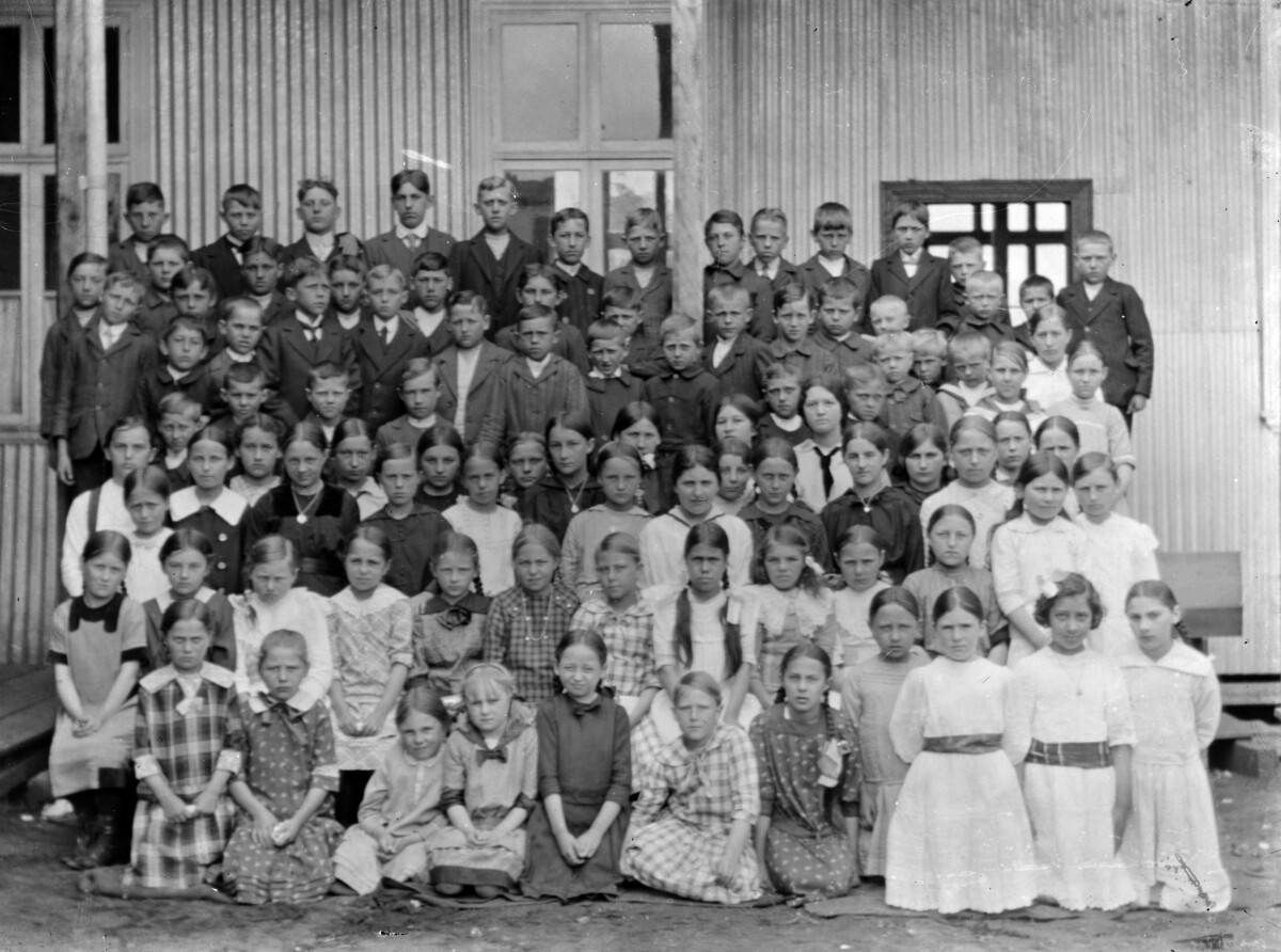 Plano general de un grupo de niños y niñas posando en la entrada de un edificio de madera y lata