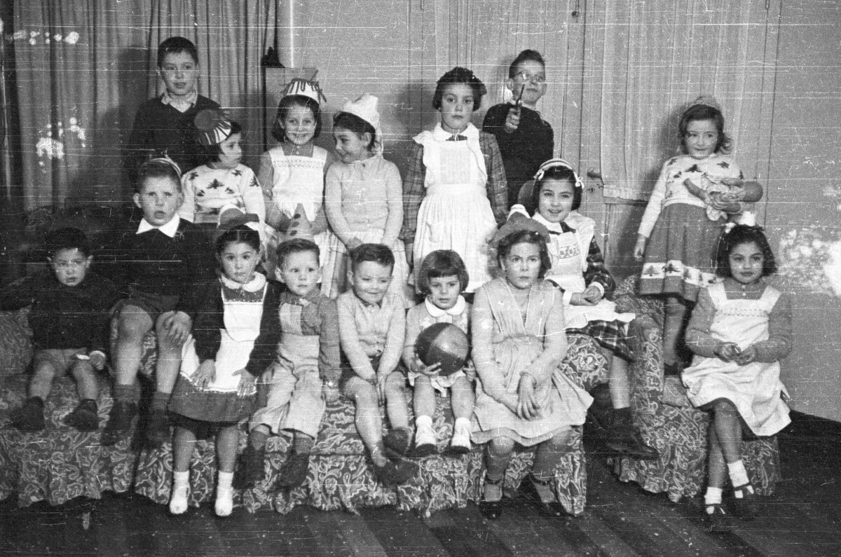 Retrato grupal de Niños y Niñas posando sentados en un sillón, la gran mayoría lleva puesto un gorro de fiesta y están en el interior de una casa