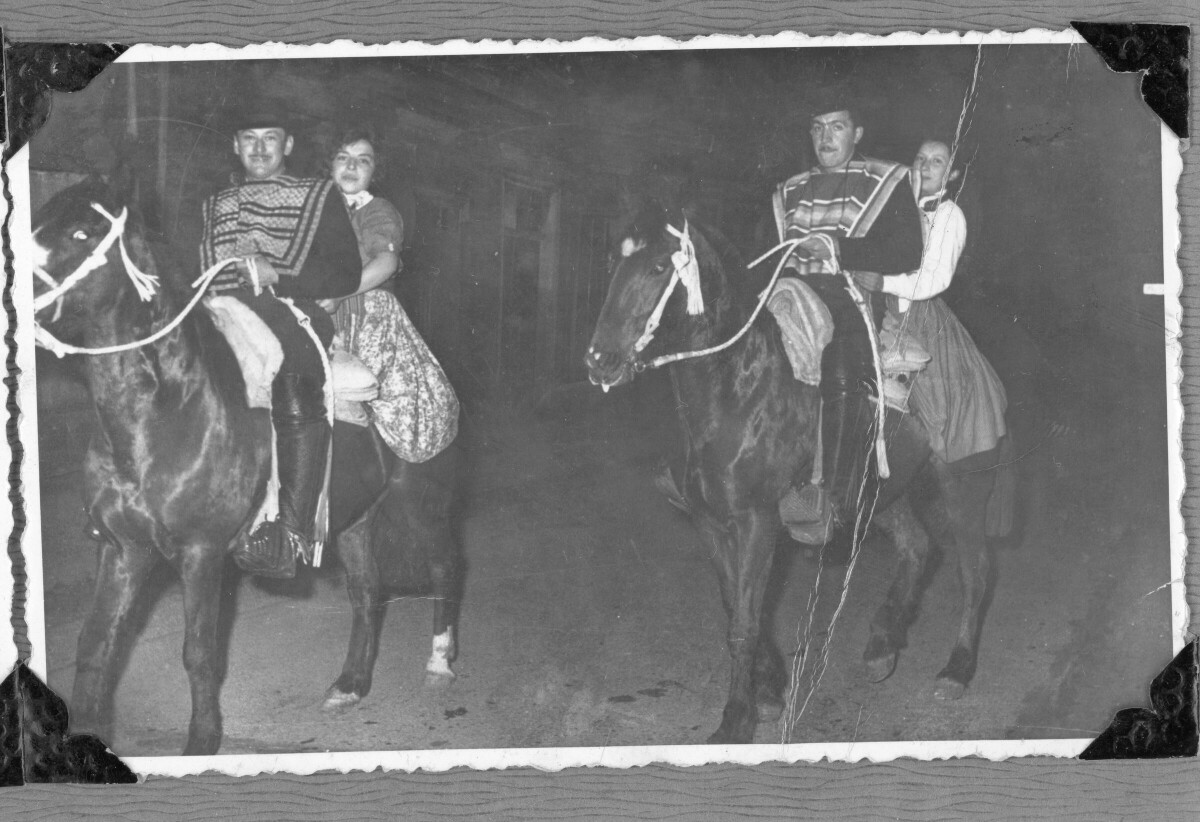 Retrato de cuatro personas montando caballos en parejas mixtas en la calle. Dos hombres están vestidos de huaso.