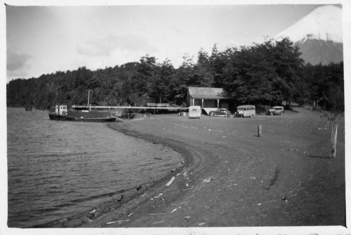 plano general de una barcaza atracada en un muelle en Petrohue. Junto a el, cuatro automóviles se encuentran estacionados, incluidos transporte de pasajeros. En el fondo se observa el volcán Osorno.