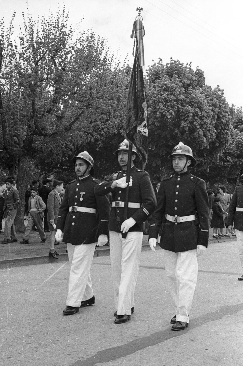 -Plano entero de tres bomberos desfilando formados en una hilera. En la hebilla de su cinturón llevan grabado el número --4--. El del medio va levantando una bandera por el mástil mientras avanzan por una calle pavimentada a un costado de la Plaza de La Concordia-
