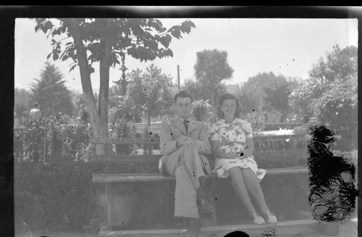 Plano medio de dos personas jóvenes (hombre y mujer) posando sentados en una banca en la plaza de alguna ciudad