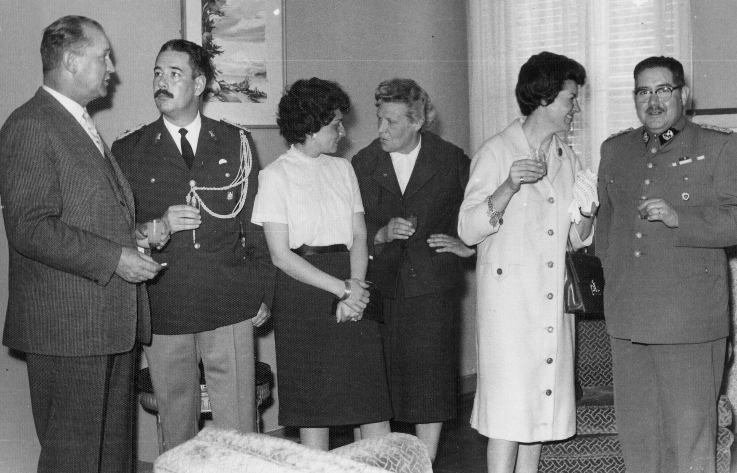 plano general de Olga Boettcher conversando con una mujer. Ambas se encuentran en el medio de la hilera, en la cual también se encuentra una mujer conversando con un carabinero y de dos hombres, de los cuales uno también es uniformado de la misma institución. Se encuentran en el interior de una infraestructura durante una celebración