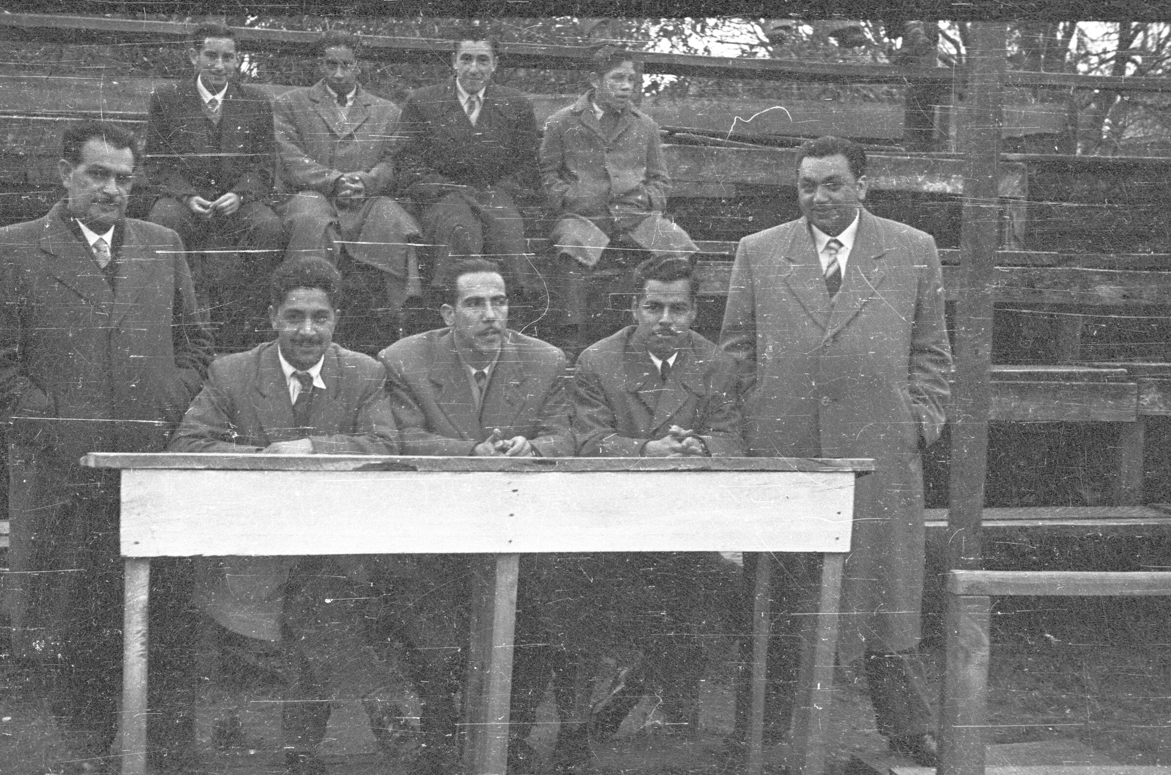 Plano entero de un grupo de hombres, jurados de un partido de baloncesto, posando sentados en una mesa, en el fondo hay dos hombres y dos niños sentados en las graderías