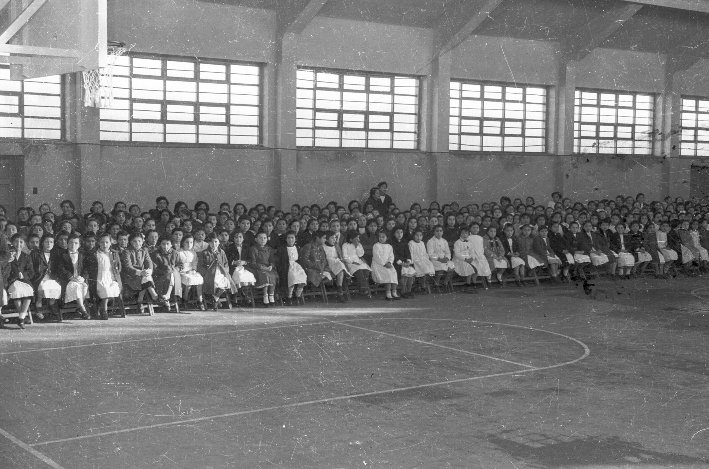 plano general de gran cantidad de niñas y Niños, algunas vestidas con delantal blanco, sentados en sillas puestas en el gimnasio de Escuela superior de niñas n°2