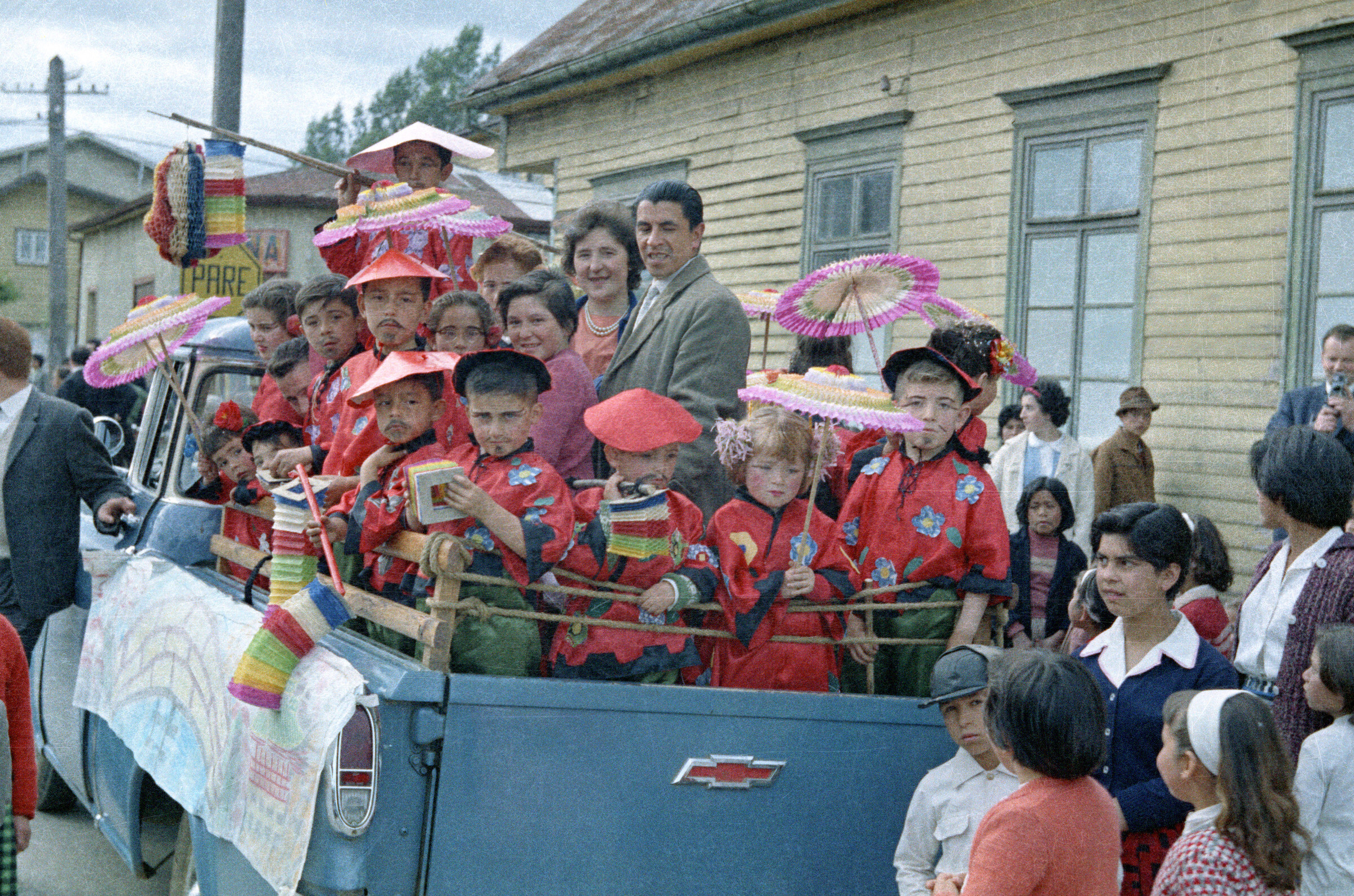 plano general de un grupo de niñas y Niños parados en la parte trasera de una camioneta, junto a Wilfred Casanova y Bella Uribe Jaramillo. Se puede observar que están disfrazados con un kimono rojo y sostienen una sombrilla durante un recorrido realizado en carros alegóricos por las calles de la Unión.