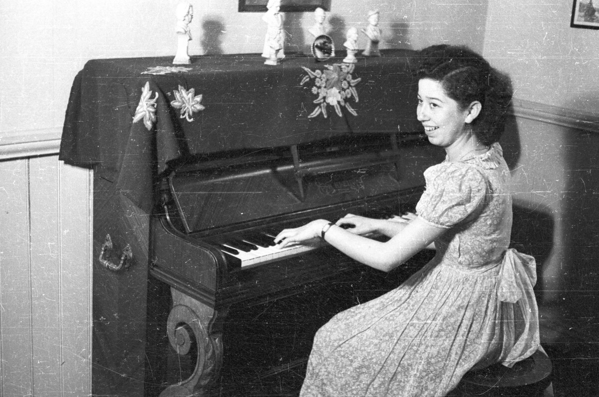 Plano entero de Norma Astudillo sentada tocando un piano de pared. Sobre el piano hay bustos de hombres en miniatura y en la pared hay colgados marcos de fotos. Está en el living de una casa.