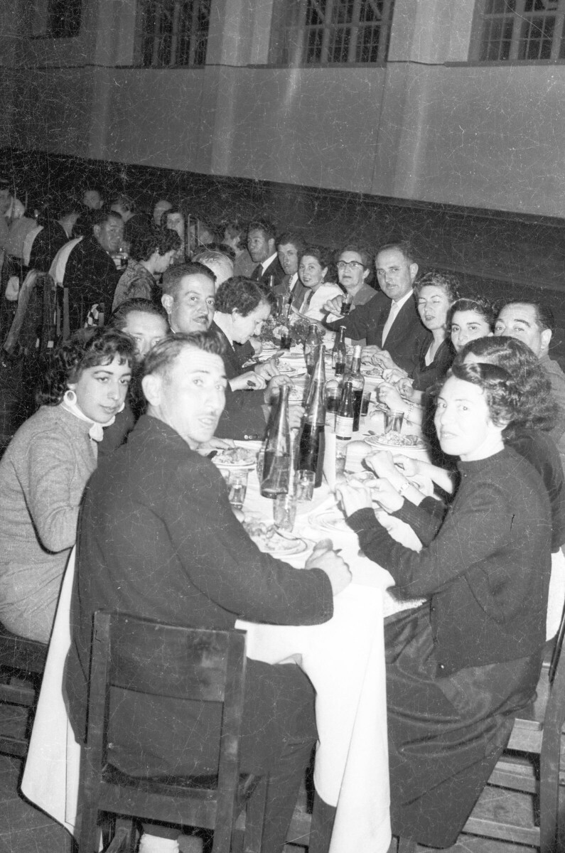 Plano entero de un grupo numeroso de Hombres y Mujeres, sentados alrededor de una larga mesa. Todos visten formalmente y algunos de ellos están mirando hacia la cámara. Se encuentran en un banquete en el gimnasio de la escuela superior de niñas Nº2