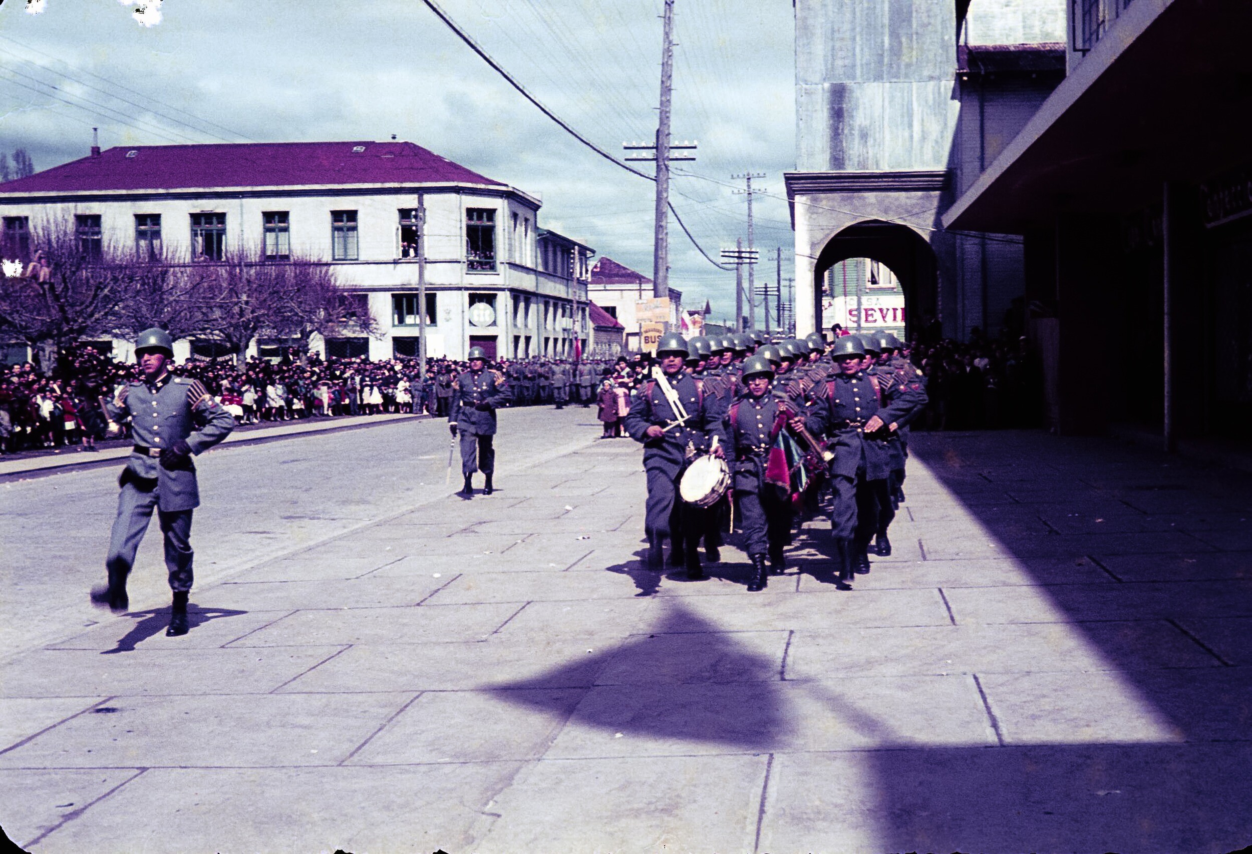 Plano entero de banda de guerra desfilando por calzada de calle Prat frente a locales comerciales de La Unión