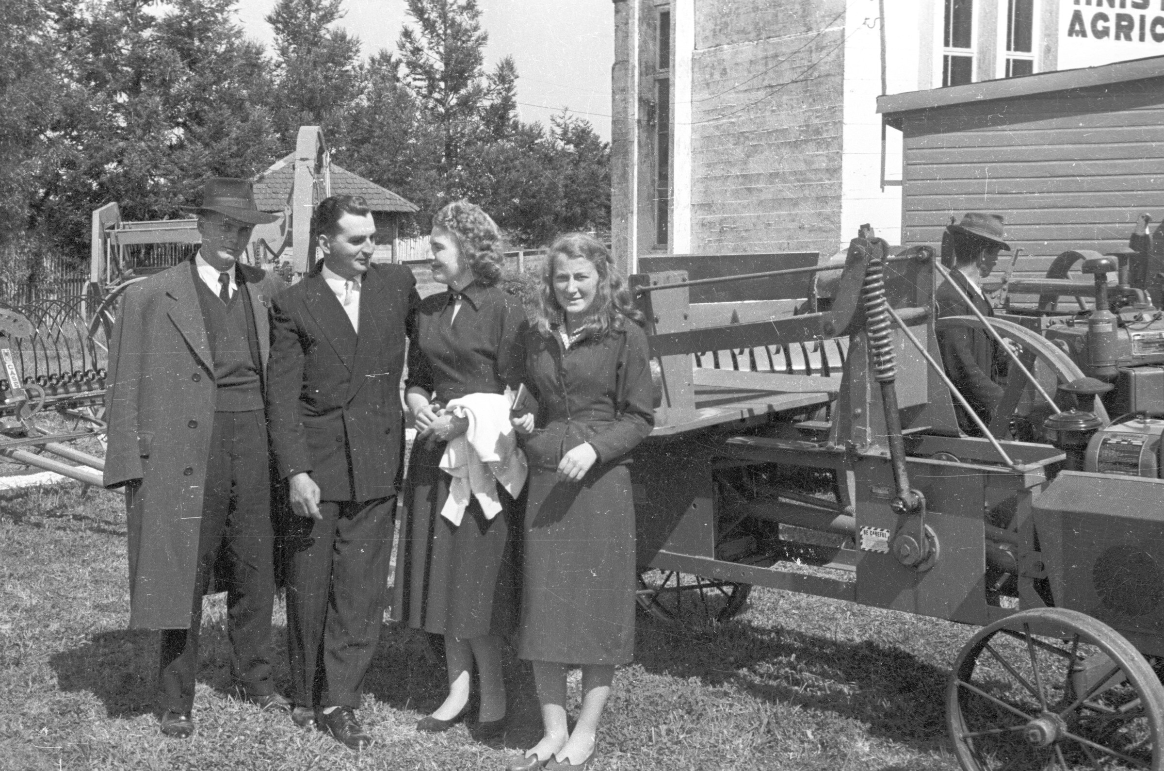 Plano entero de dos hombres vestidos de Traje y dos mujeres, parados junto a una maquinaria. El hombre que lleva sombrero y una de las mujeres se encuentran mirando hacia la cámara. Están en el interior de un recinto no identificado durante la exposición de maquinarias agroindustriales.