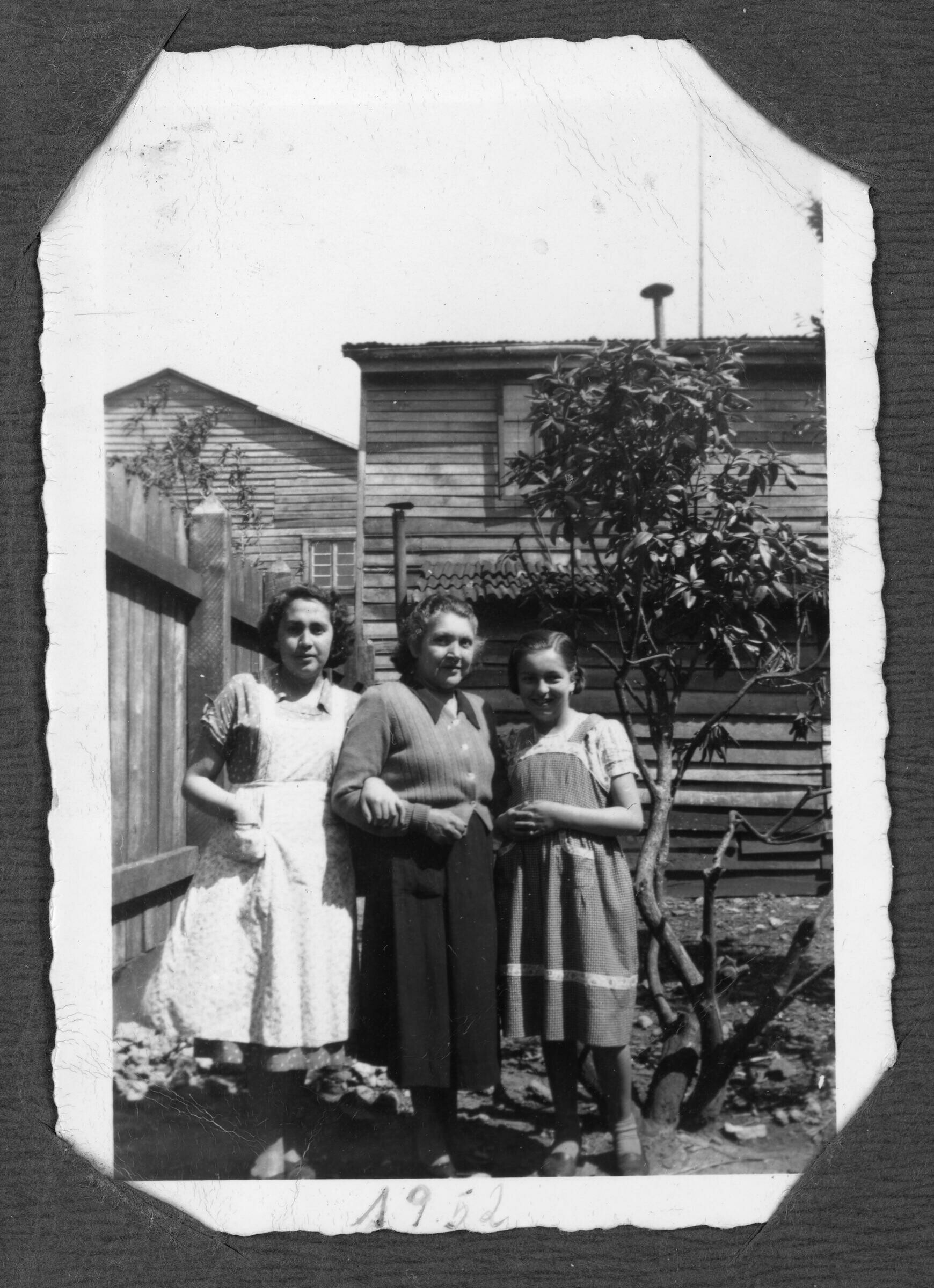 Retrato grupal de dos mujeres adultas y una niña adolescente en el patio de una casa. Están junto a un árbol y un cerco de madera. detrás hay una casa.