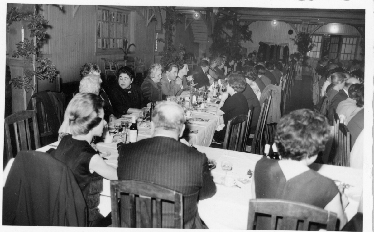 plano general de un grupo de personas sentadas alrededor de largas mesas, mientras conversan y ríen entre sí. En el fondo, hay un garzón atendiendo una mesa durante un banquete realizado en honor a Olga Boettcher tras haber finalizado su gobernación