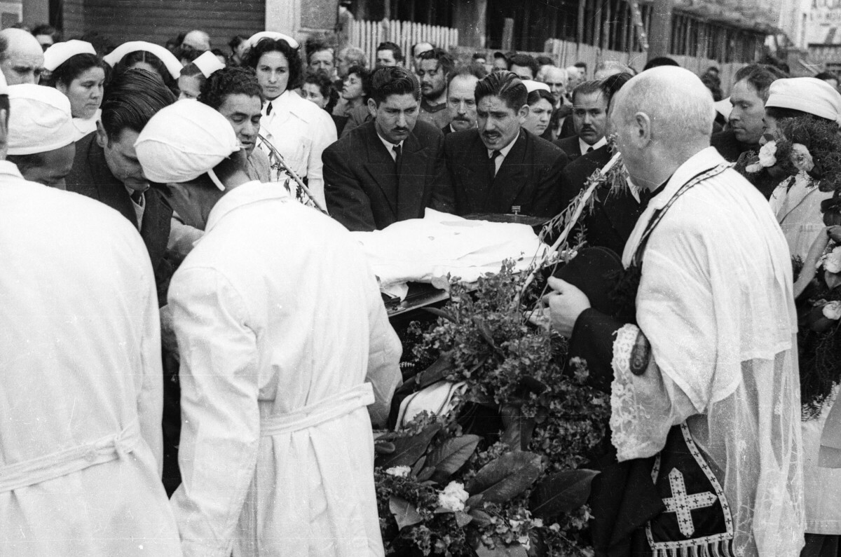 Plano entero de un sacerdote parado junto a un grupo de Hombres, alrededor del féretro de un infante. Algunos hombres van vestidos de Traje y otros van vestidos de blanco. En el fondo hay un grupo numeroso de personas que los acompañan para seguirlos hasta el cementerio ubicados en el frontis de la Parroquia San José