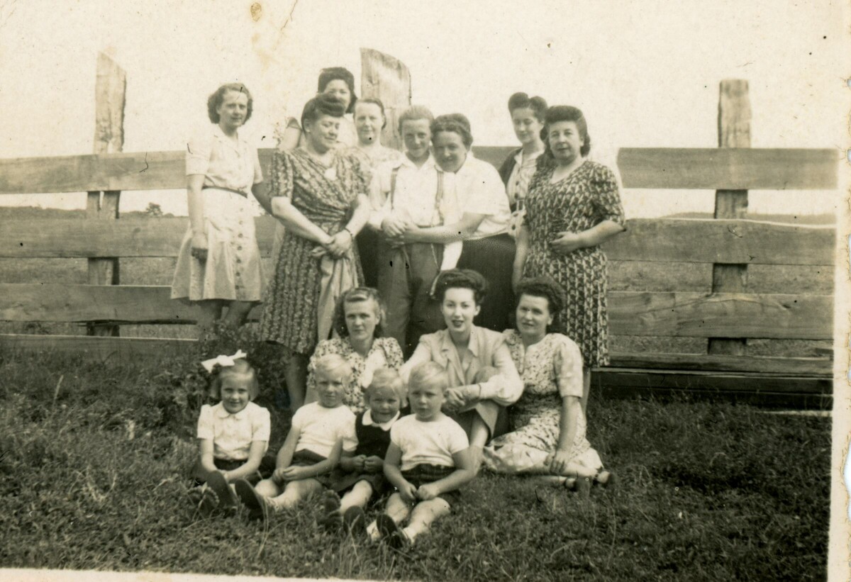 Retrato grupal de plano entero de mujeres, niñas y niños posando delante de un cerco de madera en un lugar no identificado.