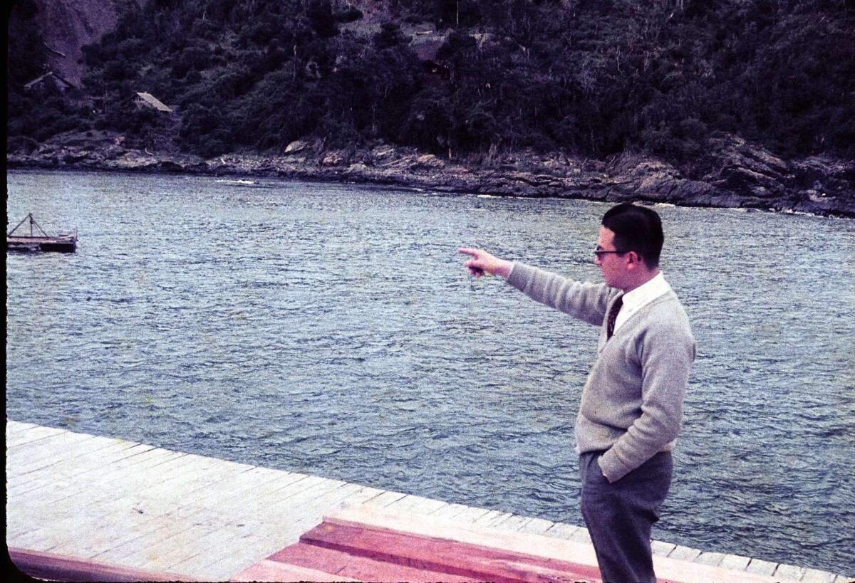 Plano entero de Hombre con Traje apuntando con el dedo en un muelle, de fondo el lago y un cerro poblado de árboles