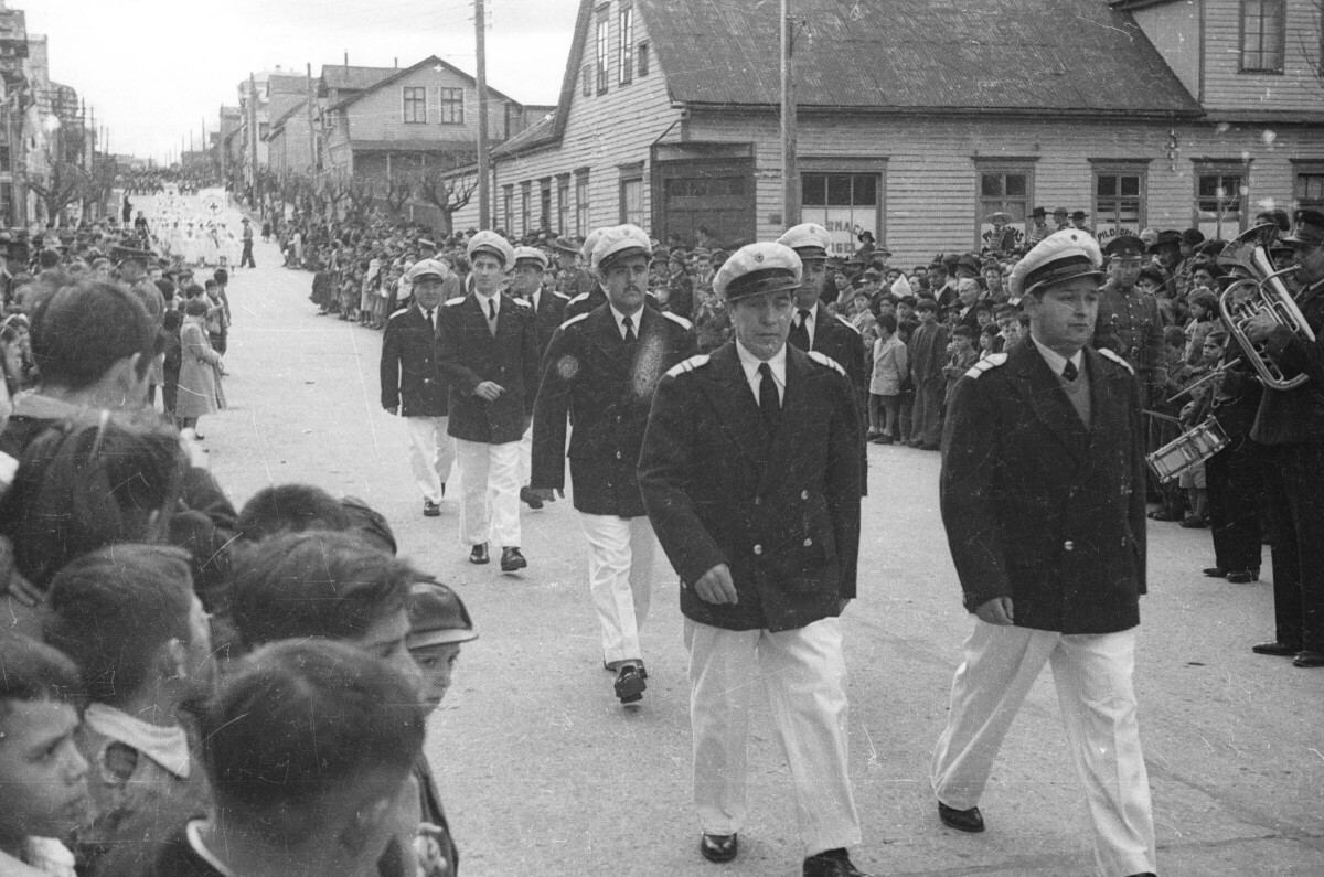 Plano entero de un grupo de Hombres uniformados, desfilando en hileras por una calle pavimentada. En ambos costados, se encuentra un grupo numeroso de Niñas, Niños, Hombres y Mujer observándolos avanzar durante el desfile realizado por una de las calles de la ciudad de La Unión