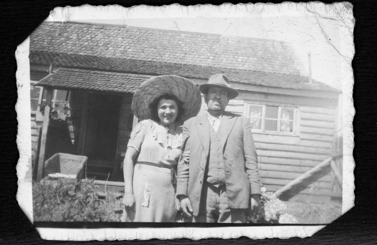 Retrato de dos personas a las afueras de una casa. La mujer viste un vestido y sombrero, y el hombre terno y sombrero