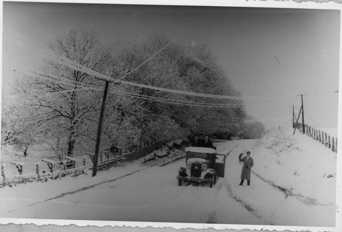 plano general cuatro hombres posando. Tres de ellos están parados en la parte trasera de un auto mientras que el cuarto está posando parado sobre la nieve. Se encuentran a la mitad de una cuesta cubierta nieve al igual que todo el lugar que los rodea. La fotografía fue tomada tras la nevazón caída en el año 1954